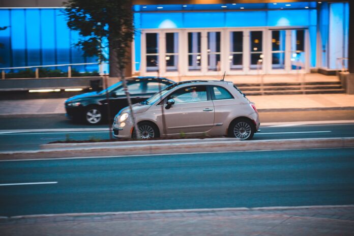 Fiat 500 jedzie nocą obok nowoczesnego, niebiesko podświetlonego biurowca Fiat 500 jedzie nocą obok nowoczesnego, niebiesko podświetlonego biurowca